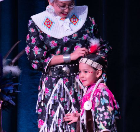 A young girl wearing a headdress and colorful clothing smiles near a women fixing her hair.