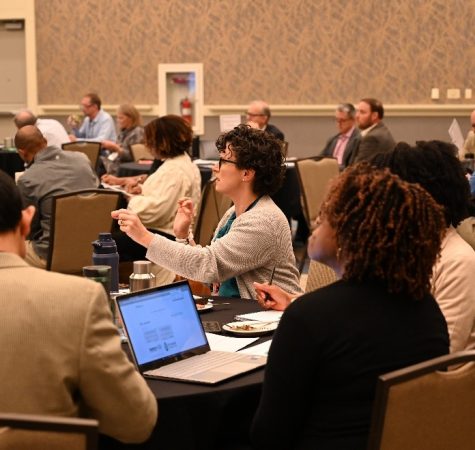 A woman with short curly dark hair speaks at a conference while sitting at a table, surrounded by other conference participants