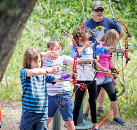 Four children practice archery outside with an adult supervising.