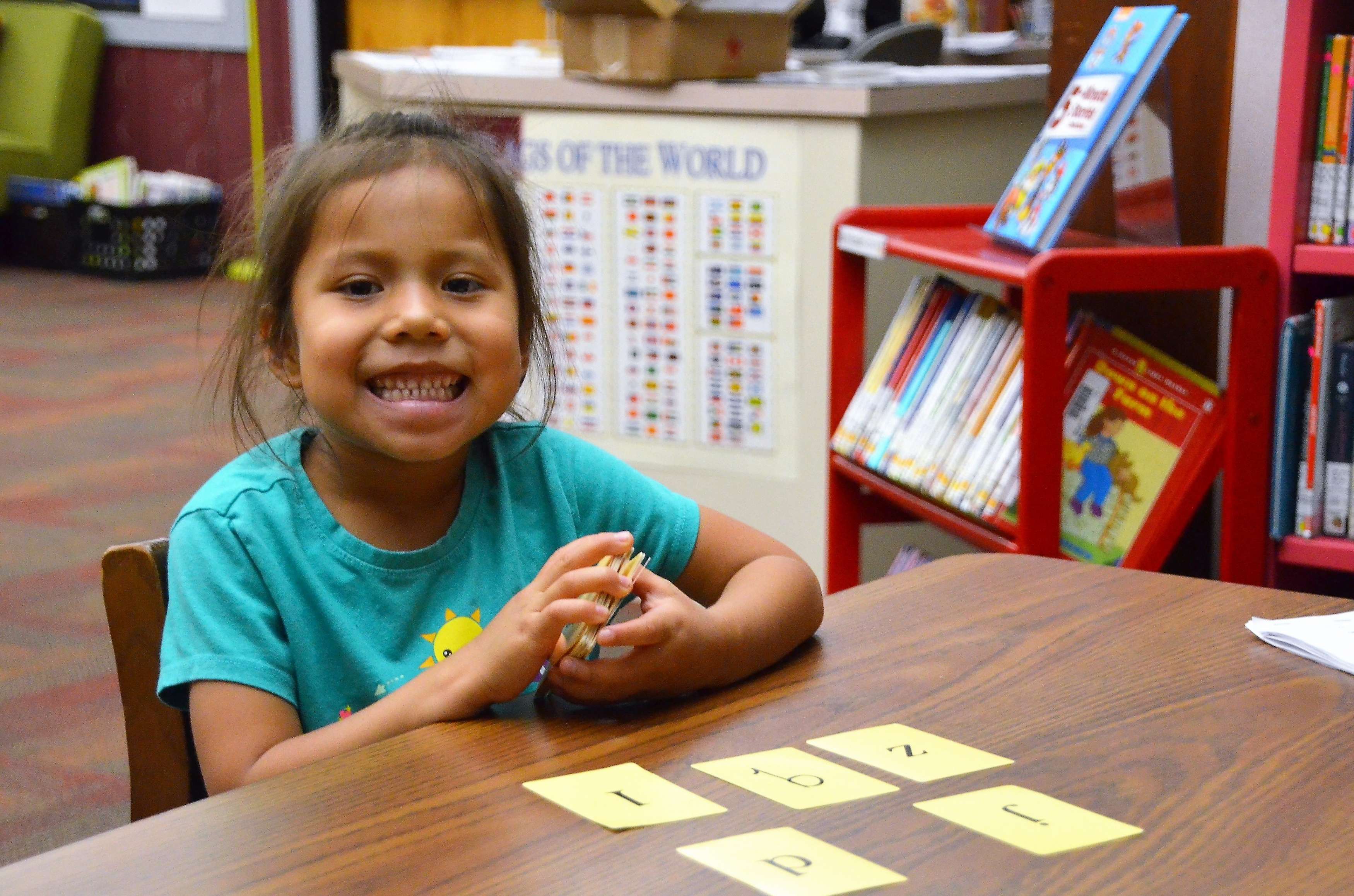 A young girl wearing a blue shirt and holding flashcards smiles for the camera.