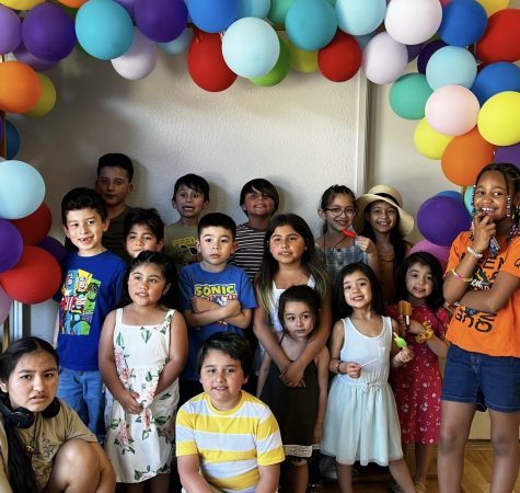 A group of children stand under a colorful balloon arch.